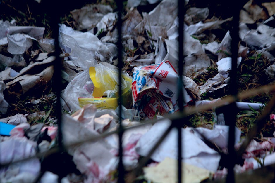 Photograph of discarded waste and litter behind a black metal fence, including crumpled paper and plastic bags, on a patch of dirt and grass. The waste appears to be illegally dumped and is surrounded