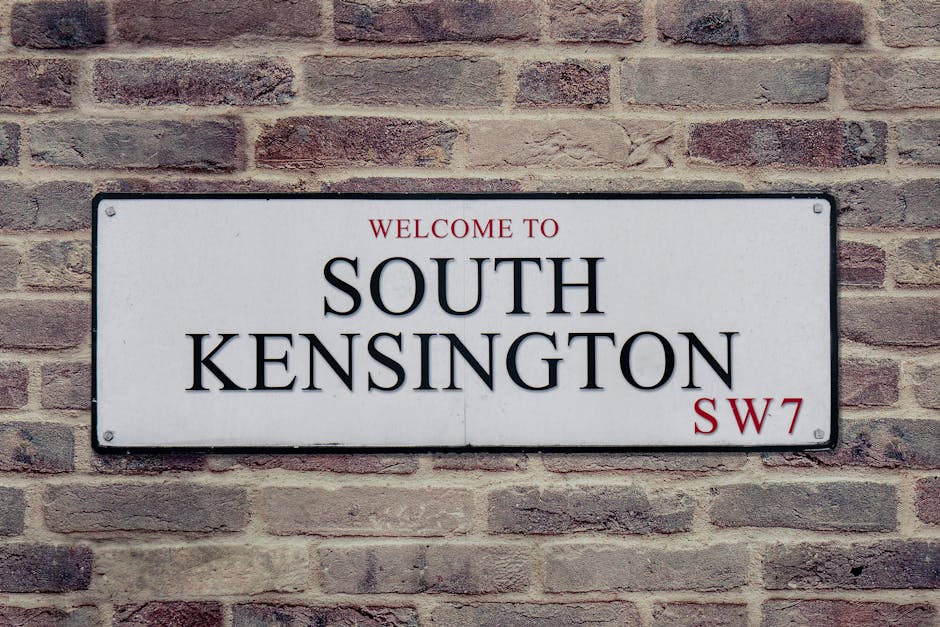 A rectangular metal street sign mounted on a brick wall, displaying the words 'Welcome to South Kensington SW7' in black and red text. The sign has a white background with black border edges, and is positioned on a red-brown brick surface with a rough texture. The lighting is even, illuminating both the sign and the brick wall, highlighting the clear and clean appearance of the sign. This image reflects the typical signage found in residential or commercial areas in South Kensington, which may benefit from surface cleaning and maintenance services offered by Kensington Cleaners as described on their website page about Kensington High Street cleaning services guide W8.