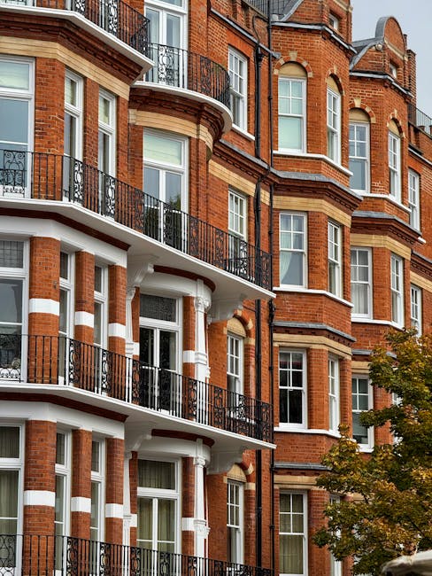 Close-up view of a multi-story residential building on Kensington High Street featuring a brick façade with decorative white accents around the windows and balconies. The balconies have black wrought-iron railings, and the windows are clean and free of dust or dirt. The image shows the exterior during daytime with natural light highlighting the building's well-maintained surface. A nearby tree with green leaves partially frames the building, emphasizing the urban setting. This scene reflects the type of surface cleaning and maintenance that Kensington Cleaners provides for residential properties, aligning with the Kensington High Street cleaning services guide W8.